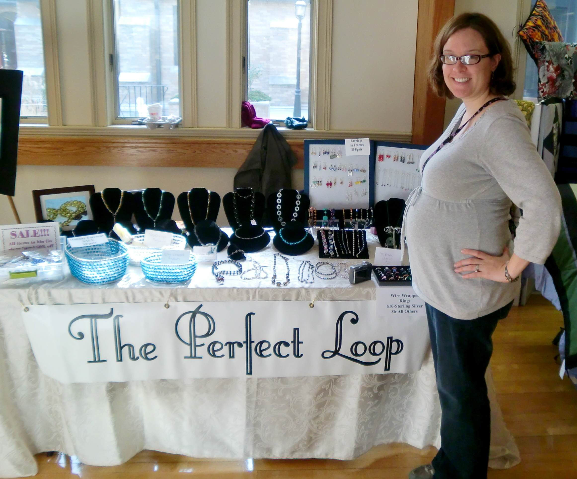 Pregnant female standing beside a craft fair vendor table featuring handmade jewelry.